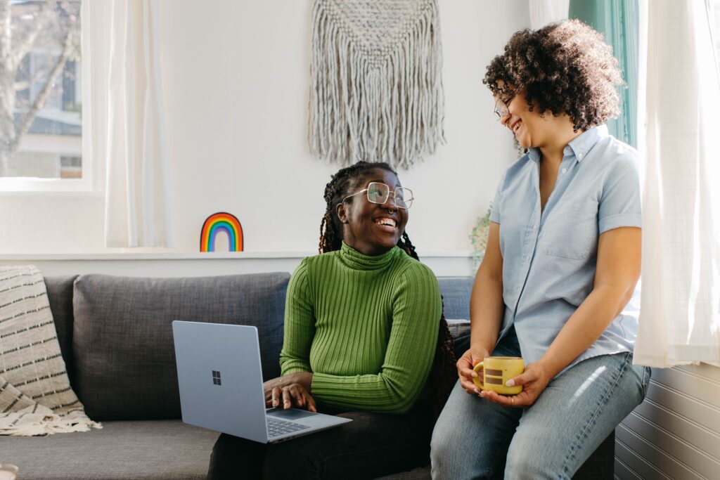 two women smiling at eachother on a sofa holding a laptop
